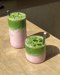 two glasses filled with green and pink drink sitting on top of a wooden table next to each other