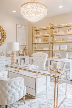 a white and gold home office with glass desk, chairs, shelves and chandelier