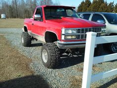 a red pick up truck parked in front of a white fence with two other trucks behind it