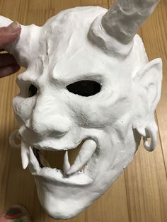 a white mask is being held up by someone's hand on a wooden table