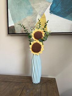 two sunflowers in a blue vase on a wooden table next to a painting