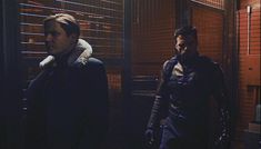 two men standing next to each other in front of a window with blinds on it