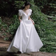 a woman in a white wedding dress standing on the edge of a river with greenery behind her