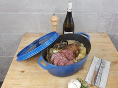 a blue pot filled with meat and vegetables on top of a wooden table next to a bottle of wine