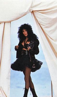 a woman in black dress and boots standing next to a white curtain with her hand on her hip