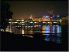 the bridge is lit up at night with lights reflecting in the water and buildings behind it