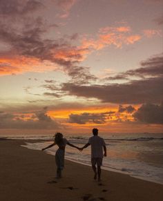 a man and woman walking on the beach holding hands as the sun sets behind them