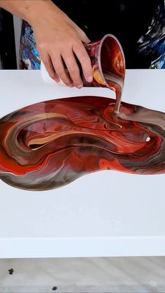 a person pouring liquid onto a piece of art that looks like a flowing red and brown swirl