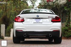 the back end of a white bmw car parked in front of a tree and shrubbery
