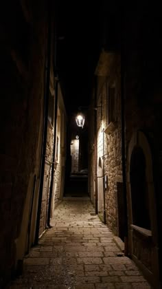 an alley way with stone walls and cobblestone pavement at night, lit by a street lamp