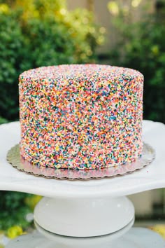 a sprinkle covered cake sitting on top of a white cake plate in front of trees