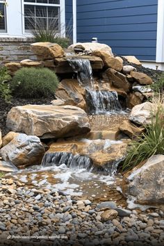 a small waterfall in the middle of a garden with rocks and plants around it, next to a blue house