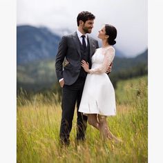 a man and woman standing next to each other in tall grass with mountains in the background