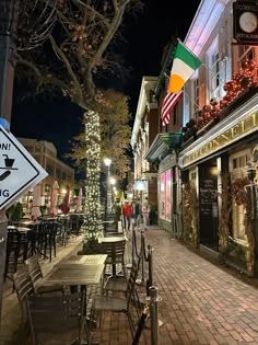 a city street with tables and chairs on the side walk at night, lit up by christmas lights
