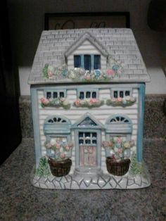a white and blue ceramic house with flowers on the windowsill, sitting on a granite countertop