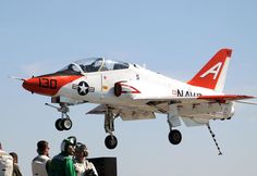 a red and white fighter jet flying through the air with people looking on from below