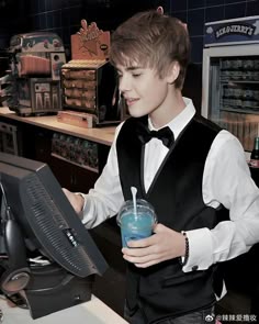 a young man wearing a vest and holding a drink in front of a computer screen