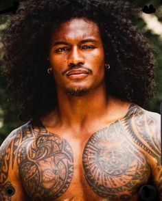 a man with tattoos on his chest standing in front of trees and looking at the camera