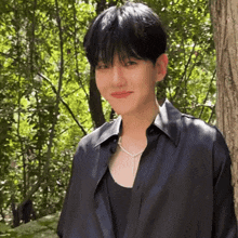 a young man wearing a black shirt and a necklace is standing next to a tree in the woods .