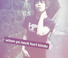 a young man in a black t-shirt is standing in front of a wall holding his neck .