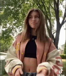 a young woman wearing a pink varsity jacket and a black crop top is standing in front of a tree .