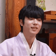 a young man wearing a white kimono is sitting in front of a wooden wall and smiling .