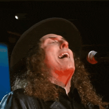 a man with long curly hair is singing into a microphone while wearing a black hat