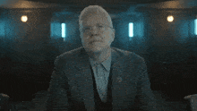 an older man in a suit and tie is sitting with his arms crossed in a dark room .
