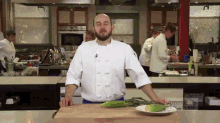 a man in a chef 's uniform stands in a kitchen with a plate of corn on the cob