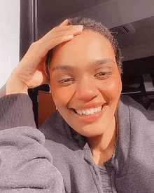 a woman wearing a grey sweatshirt is smiling with her hand on her head