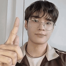 a young man wearing glasses and a brown jacket is giving a thumbs up sign .