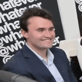 a man in a suit is smiling while sitting in front of a sign that says `` what do you hate ? ''