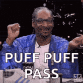 snoop dogg is sitting at a table with his fist in the air and the words puff puff pass written on the screen .