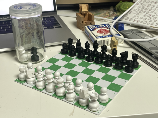 A physical chessboard. The board is white and green with notions. The pieces are white and black and short in height.